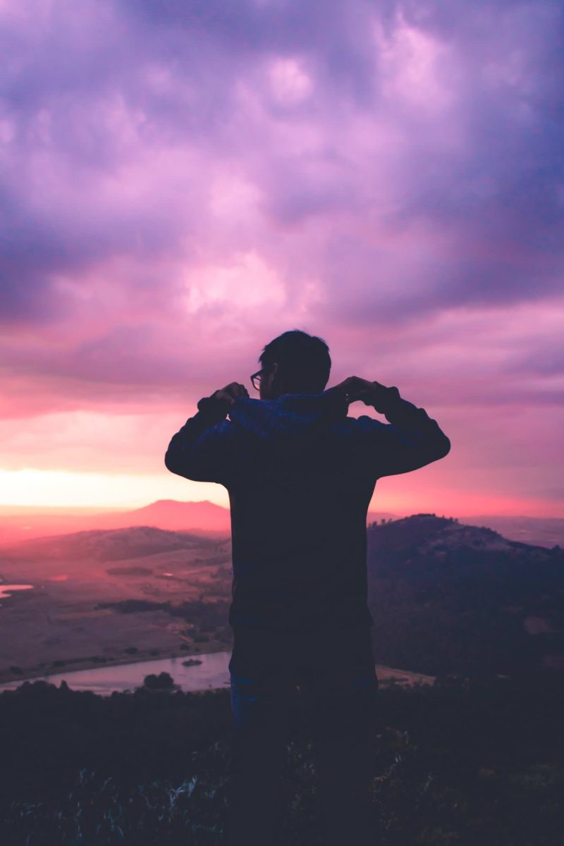 man trying to put on his hood, standing on top of a mountain, in front of colorful beautiful sunset