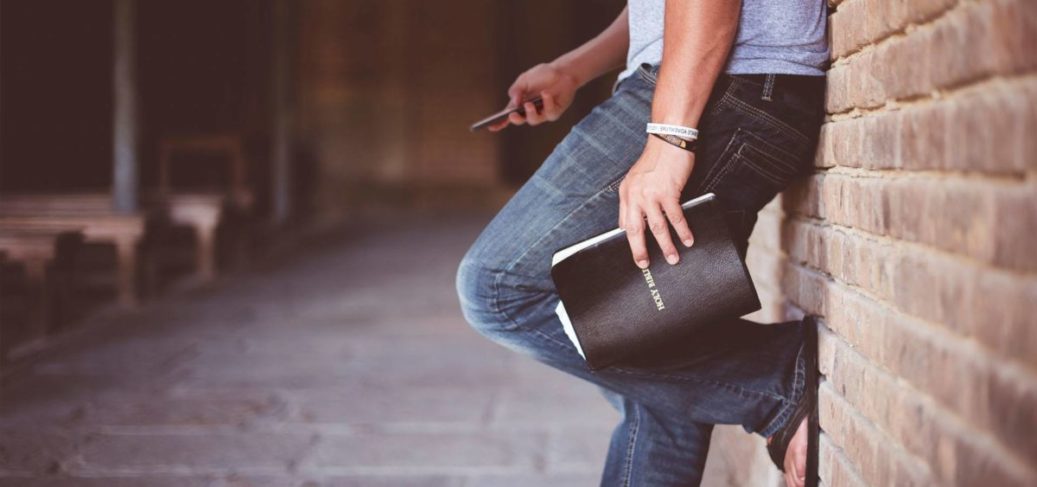 person leaning on brick wall with foot up, bible and phone in hands