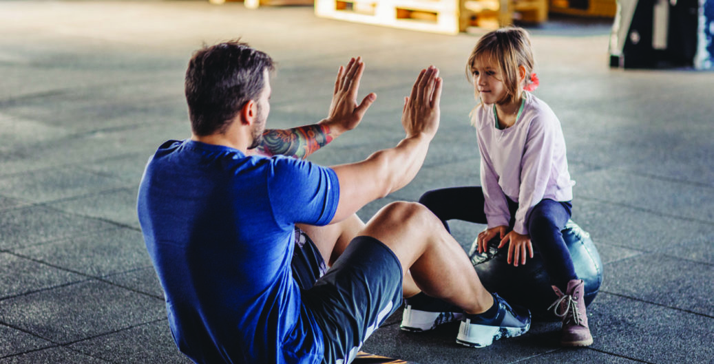 Father doing his training in gym while his little daughter supporting him.