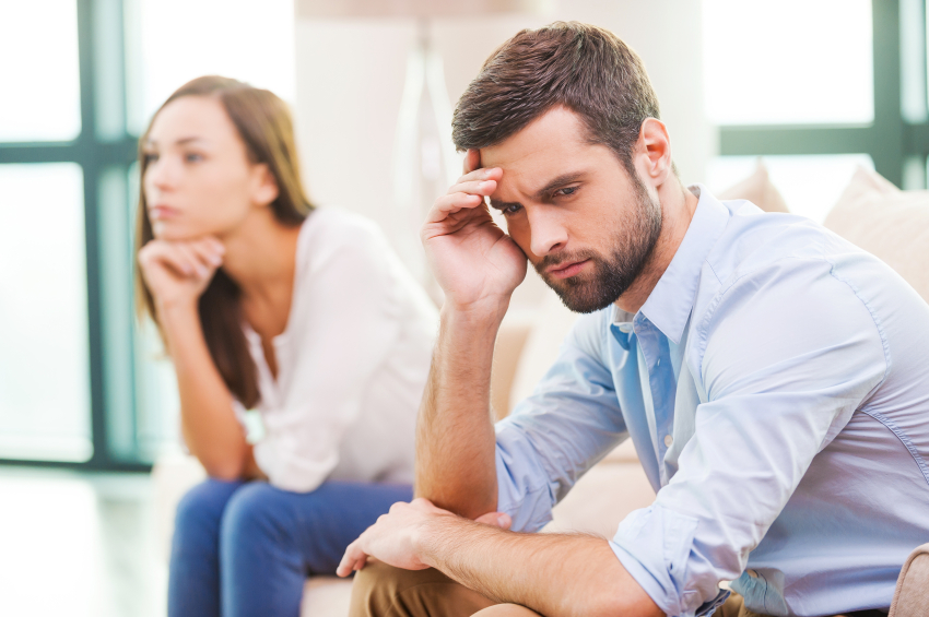 Depressed young man holding hand on head and looking away while woman sitting behind him on the couch