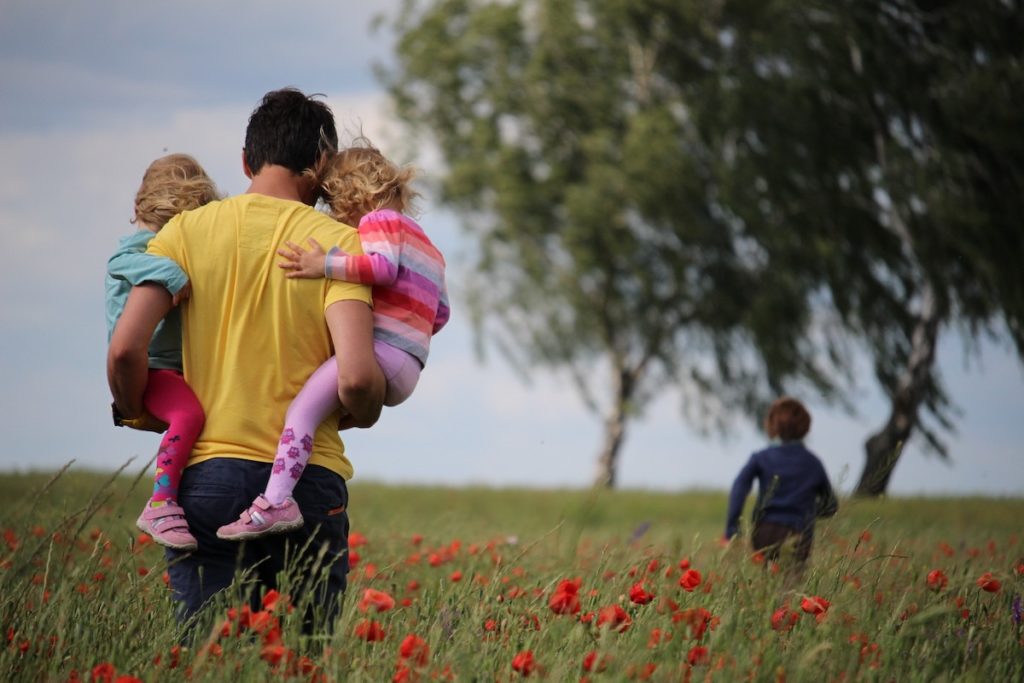 father holding 2 kids and other kid running in field