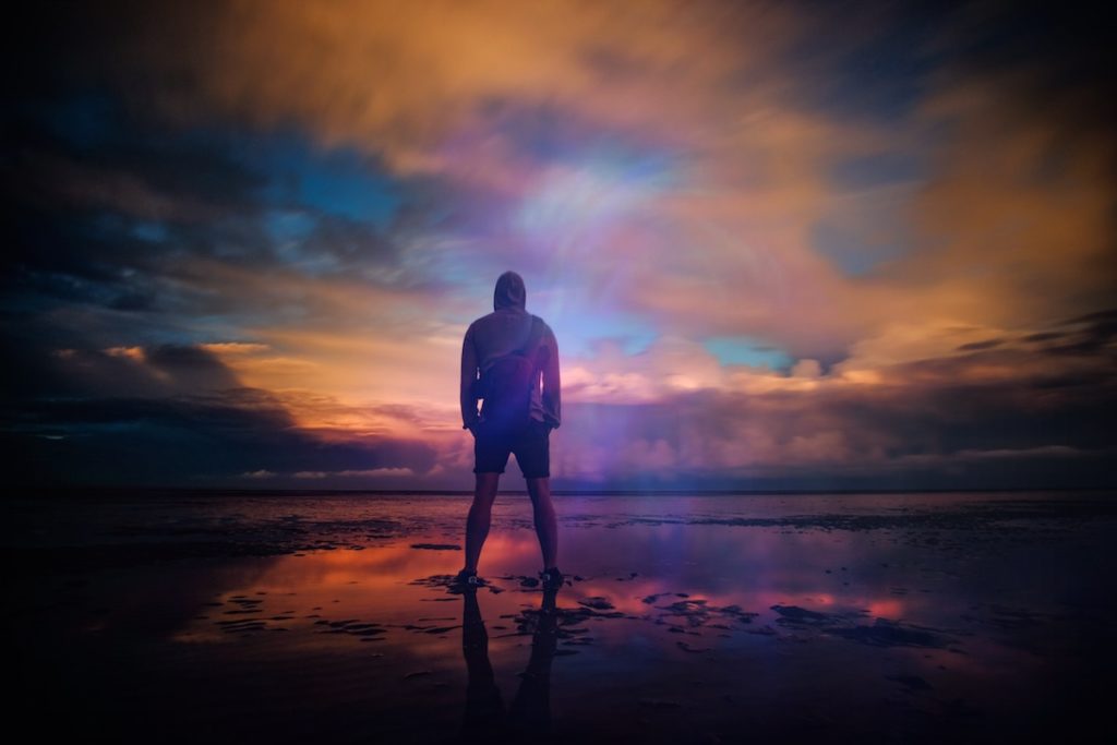 man standing on beach with multicolored sunset