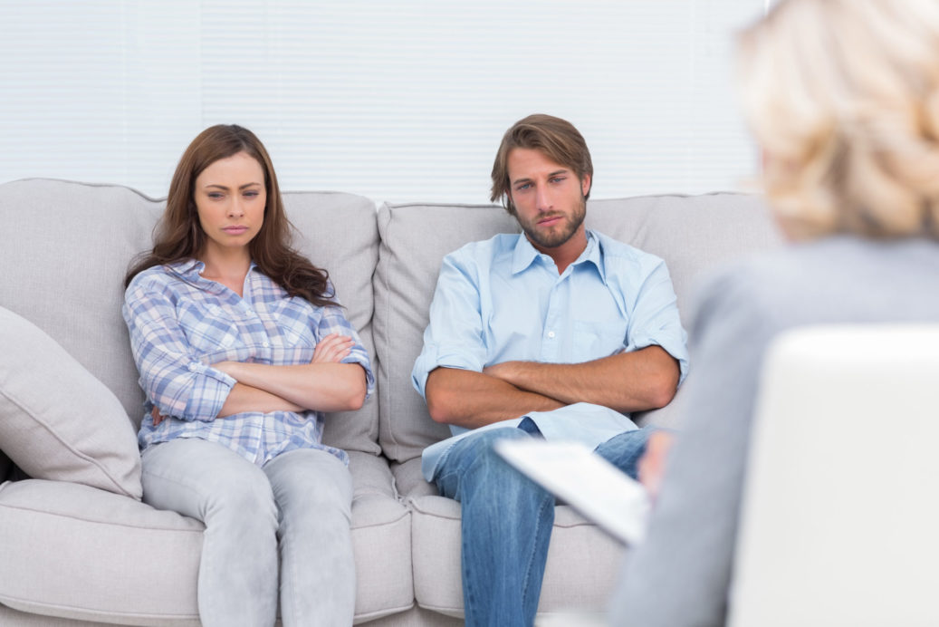 Upset couple sit on a sofa with arms crossed during a psychotherapy