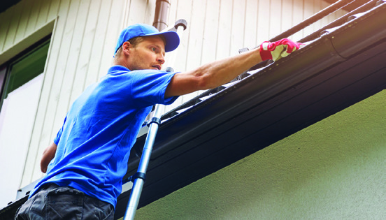 man on ladder cleaning house gutter from leaves and dirt