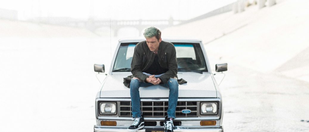 man with blue hair sitting on hood of an old white car sad