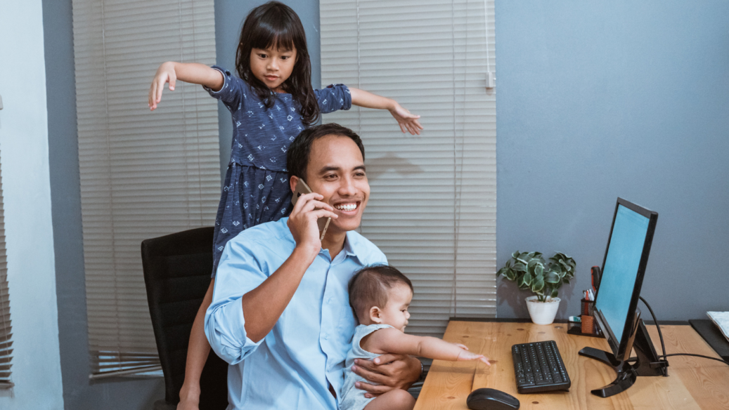 dad busting working at home while holding a baby on his lap and his daughter dancing on his chair