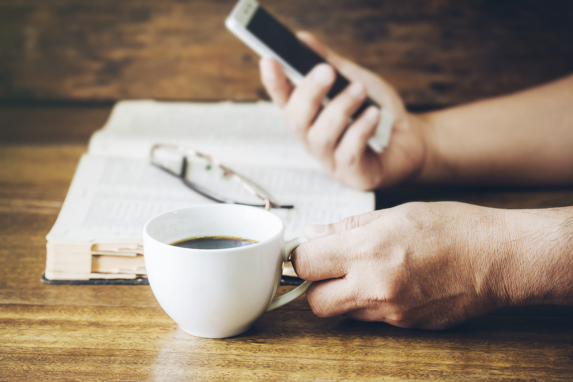 man hand holding a cup of coffee and smart phone over holy bible on wooden background