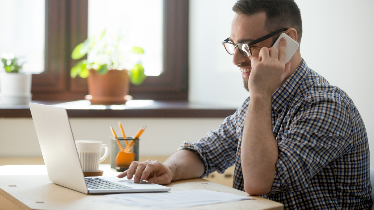 Smiling male worker talking over cell, consulting client online, typing message on laptop, writing email, giving help, assistance. Freelancer person speaking on phone, working. Concept of multitasking